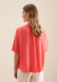 Blusa corallo realizzata in tessuto leggero, caratterizzata da una vestibilità ampia, maniche corte e un orlo arrotondato, abbinata a pantaloni chiari.