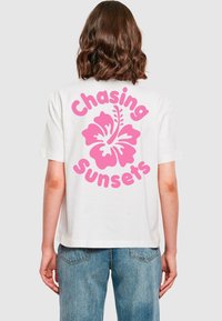 Bijela pamučna majica s velikim grafičkim motivom ružičastog hibiskusa i tekstom "Chasing Sunsets" otisnutim velikim ružičastim slovima na leđima.