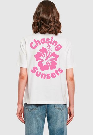 T-shirt en coton blanc avec un grand graphique d'une fleur d'hibiscus rose et le texte "Chasing Sunsets" imprimé en lettres roses en gras au dos.