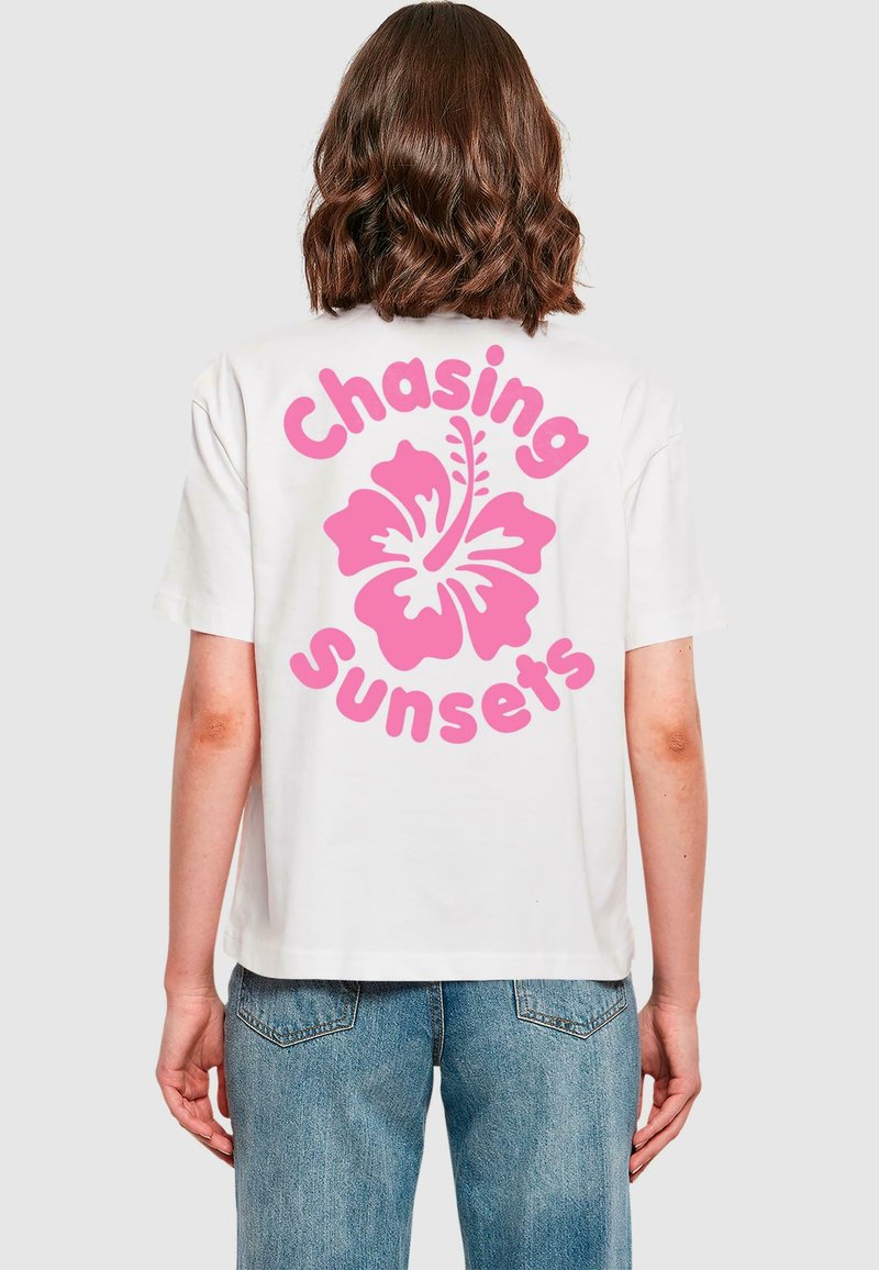 Bijela pamučna majica s velikim grafičkim motivom ružičastog hibiskusa i tekstom "Chasing Sunsets" otisnutim velikim ružičastim slovima na leđima.