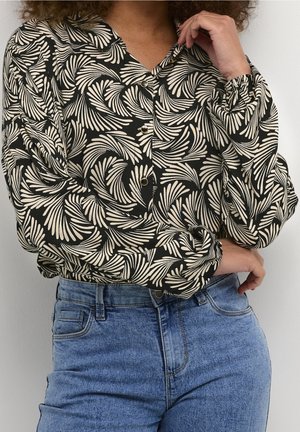 Kvinde iført en sort knapbluse med hvide snurrende bladmønstre og højtaljede blå jeans, armene krydsede og hånden rører hagen.