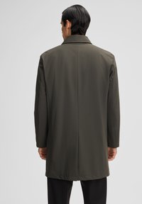Lungo cappotto verde oliva con una texture liscia, colletto classico e taglio dritto, caratterizzato da una fessura sul retro per facilitare i movimenti.