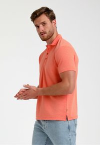 Koralen kratek polo s kratkimi rokavi, izdelan iz teksturirane tkanine, s trojnim gumbnim zapenjanjem in stranskimi odprtinami, v kombinaciji s svetlo modrimi kavbojkami.