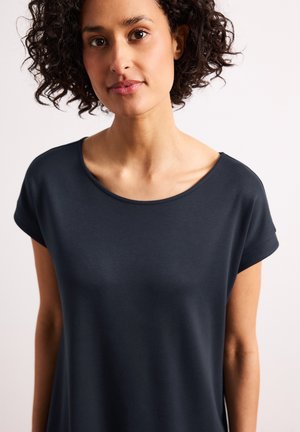 Femme aux cheveux bouclés foncés portant un haut bleu marine à manches courtes, face à l'objectif sur un fond clair uni.