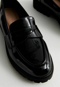 Mocassins en cuir verni noir avec une finition lisse, dotés d'une sangle décorative sur le dessus et d'une semelle en caoutchouc texturé.