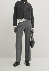 Schwarze kurze Jacke mit zwei vorderen Taschen, kombiniert mit grauen, gestreiften weiten Hosen. Trägt schwarze Schuhe und hält eine kleine schwarze Tasche.