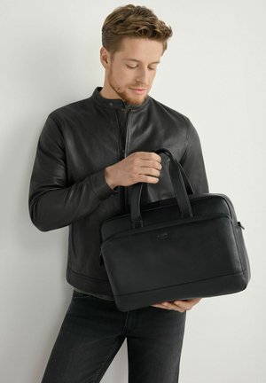 Mężczyzna w czarnej skórzanej kurtce trzyma i patrzy na czarną skórzaną teczkę na jednolitym białym tle.