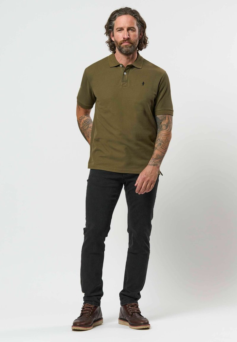Homme aux longs cheveux et tatouages porte un polo vert olive, un jean noir et des bottes marron, debout devant un fond uni clair.