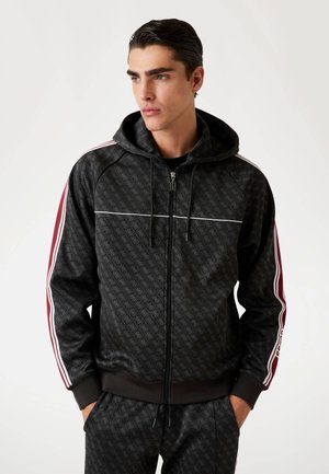 Sudadera con cremallera negra con un patrón de logotipo repetido, que presenta franjas color burdeos y blancas en las mangas. Confeccionada en una tela suave y texturizada.