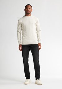 Pulover tricotat de culoare bej deschis, cu mâneci lungi, asortat cu blugi închis la culoare și teniși albi. Puloverul are un guler rotund și manșete cu bordură.