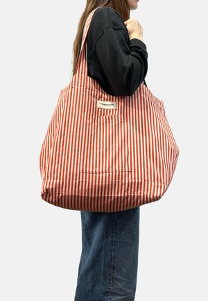 Personne portant un pull noir et un jean bleu tient un grand sac fourre-tout rayé rouge et blanc avec l'étiquette « rivedroite » sur l’épaule.