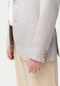 Blazer gris clair en tissu lisse, avec un col cranté, une poche unique et cinq boutons beiges sur le poignet. Coupe élégante et ajustée.