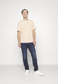 T-shirt a righe in crema pallido con strisce orizzontali arancioni, abbinato a jeans blu scuro e sneakers bianche. Taglio semplice e casual.
