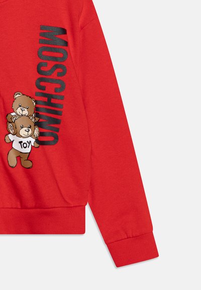 Rød hettegenser med ribbestrikket nederkant og ermer. Har et stort grafisk bilde av to brune kosedyrbjørner og "MOSCHINO" i fet svart skrift.
