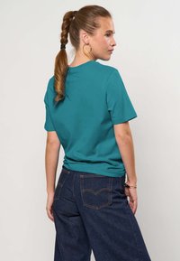 Femme aux cheveux tressés portant un t-shirt bleu sarcelle à manches courtes et un jean bleu foncé, debout de profil sur un fond clair uni.