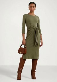 Vestido justo verde oliva com mangas de três quartos, apresentando um detalhe de laço na frente. Combinado com botas castanhas até ao joelho e uma pequena bolsa castanha.