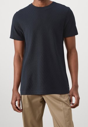 Granatowa teksturowana koszulka z okrągłym dekoltem, krótkim rękawem i subtelnym wzorem tkanym. Połączona z beżowymi spodniami.