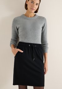 Mujer vestida con un suéter gris de canalé y una falda negra con cordón y bolsillos, de pie contra un fondo de pared beige.