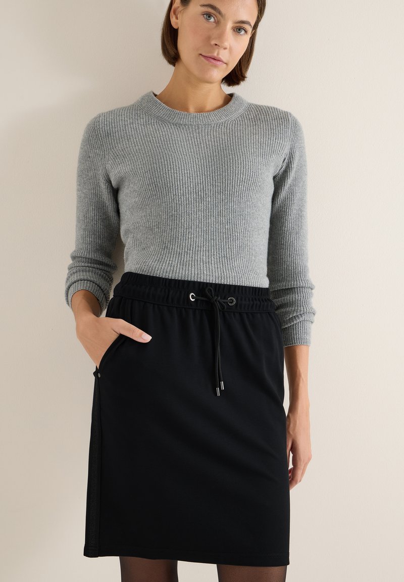 Mujer vestida con un suéter gris de canalé y una falda negra con cordón y bolsillos, de pie contra un fondo de pared beige.