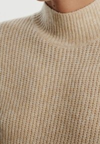 Pull beige avec une texture côtelée et un col haut ajusté. Le matériau semble doux et chaud, avec des variations subtiles d'intensité de couleur.