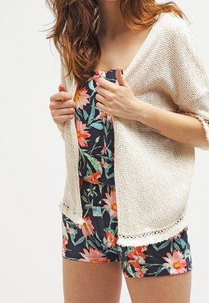 Frau trägt einen marineblauen Overall mit Blumenmuster und hält die Vorderkanten einer cremefarbenen Strickjacke.
