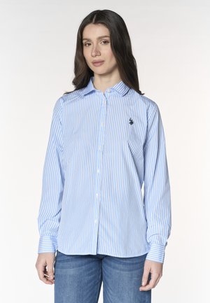 Giovane donna che indossa una camicia a righe blu e bianche con bottoni e jeans blu, in piedi su uno sfondo chiaro.