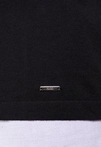 Tessuto a maglia nero con un piccolo tag rettangolare in metallo argentato etichettato "HUGO" cucito vicino al bordo inferiore.