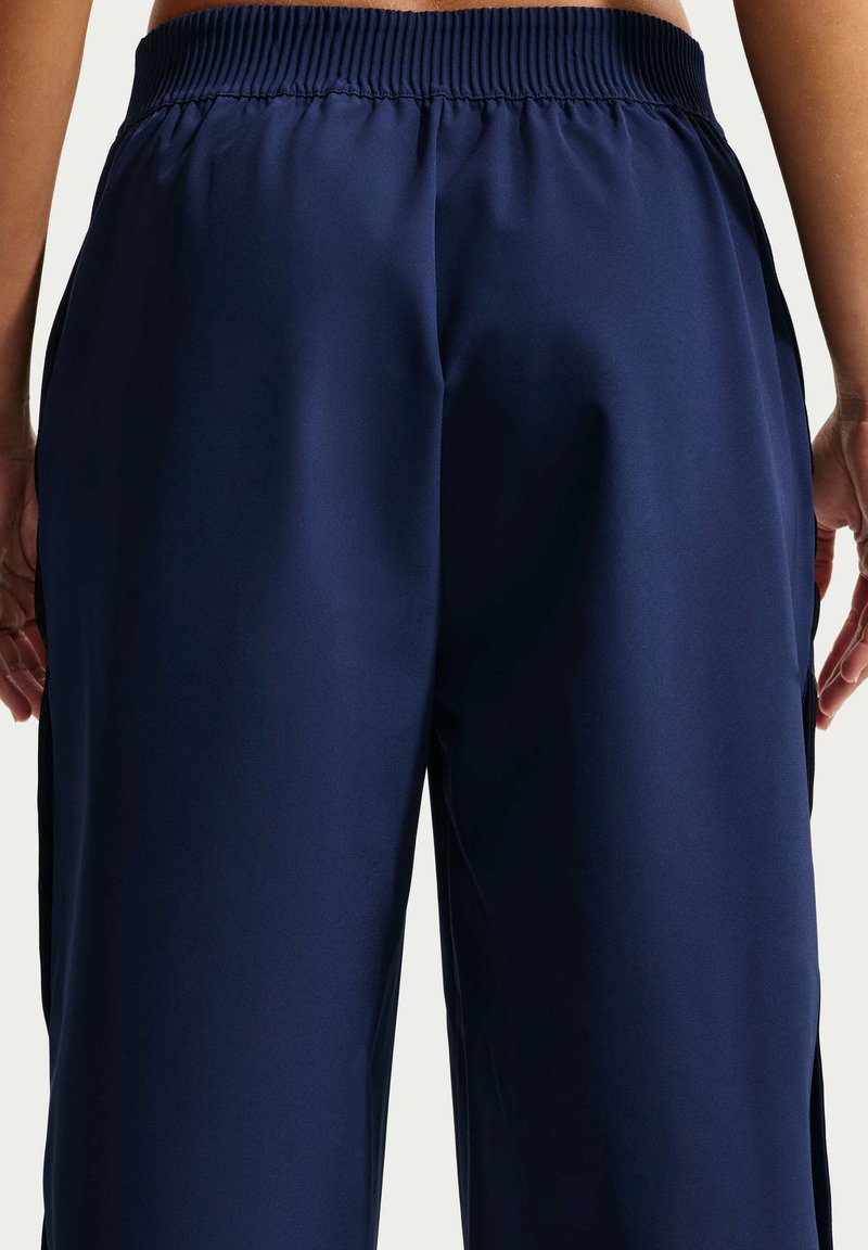 Rückansicht einer Person, die locker sitzende marineblaue Hosen mit elastischem Bund trägt, Hände entspannt an den Seiten, vor hellem Hintergrund.