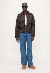 Veste matelassée brun avec un col brun, crop top blanc, jeans bleu clair à jambes larges et bottes compensées beiges ; assortie à des lunettes de soleil noires.