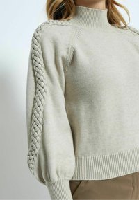 Ljusgrå tröja med hög hals, texturerad flätad detalj på ärmarna samt ribbad mudd och nederkant. Mjuk stickad material.