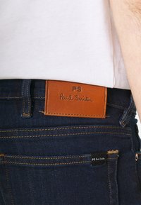 Tmavě modré džínové kalhoty s hnědým koženým štítkem označeným "PS Paul Smith", s oranžovým prošitím a malou černou nášivkou na kapse.