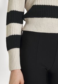 Pull tricoté avec des rayures horizontales crème et noires, tissu texturé, poignets et ourlet côtelés, associé à un pantalon noir sleek.
