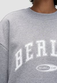 Szary sweter z okrągłym dekoltem, z białym, zniszczonym nadrukiem z napisem "BERLIN" oraz akcentem logo poniżej. Miękka, teksturowana tkanina.