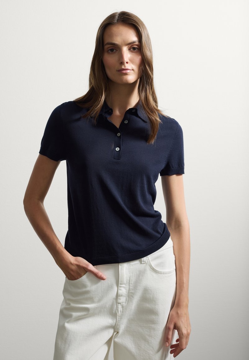 Polo shirt blu navy con maniche corte, colletto a costine, pattina con tre bottoni e un design aderente, abbinato a pantaloni bianchi.