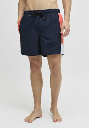 Man wearing navy blue swim shorts with coral and white side panels, standing barefoot against a plain light background.