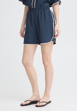 Kvinde iført marineblå shorts med elastisk talje og hvidt bølget kantbånd, parret med sorte klipklappere, stående foran en hvid baggrund.