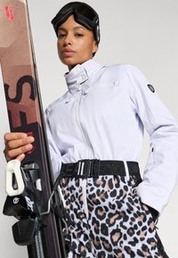 Jachetă de schi albă cu guler înalt, curea neagră și pantaloni de schi cu imprimeu leopard. Schiuri ținute într-o mână, ochelari de ski pe cap.