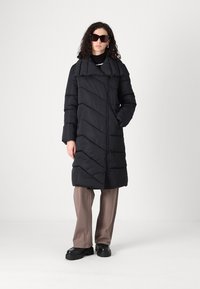 Melnā pūkainā jaka ar chevron šuvēm, liels apkakle, priekšējais rāvējslēdzis, valkāta virs melnas turtlenecka, apvienota ar bežām biksēm un masīvām melnām kurpēm.