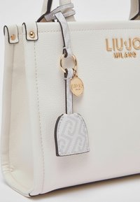 Sac à main en cuir blanc texturé avec quincaillerie dorée, comprenant un charm gris marqué et un design structuré avec un bord noir contrastant.