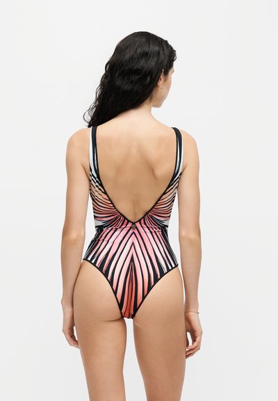 Femme aux longs cheveux foncés portant un maillot de bain une pièce à rayures roses, noires et blanches, vue de dos sur un fond uni.