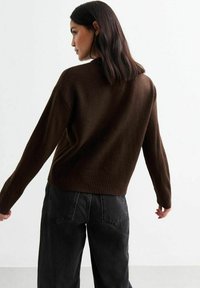 Kvinna med axellångt mörkt hår iförd en mörkbrun tröja och svarta jeans, vänd något åt sidan mot en enfärgad bakgrund.