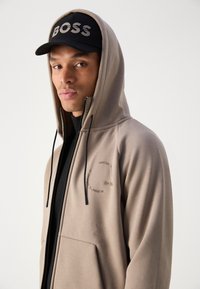Haut de survêtement beige zippé avec une capuche à cordon, arborant un logo et un texte sur la poitrine. Associé à une casquette noire affichant le mot "BOSS."