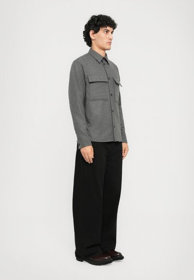 Gray button-up shirt with two front pockets, paired with loose-fitting black trousers. The shirt features a collar and long sleeves.
