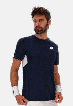 Hombre con barba que lleva una camiseta deportiva azul marino y muñequeras blancas, de pie frente a un fondo claro y liso.