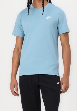 Person trägt hellblaues Nike-T-Shirt und schwarze Nike-Hose, steht mit entspannten Armen vor einem einfarbigen weißen Hintergrund.