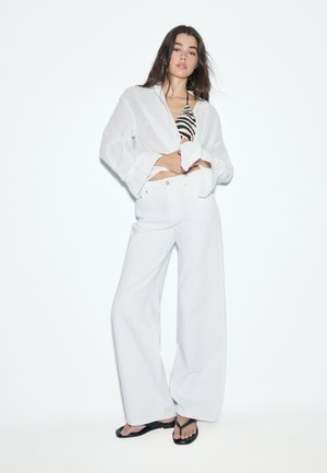 Donna che indossa pantaloni bianchi a gamba larga, camicia bianca trasparente oversize sopra un top bikini con stampa zebrata e infradito nere, in piedi su sfondo bianco.