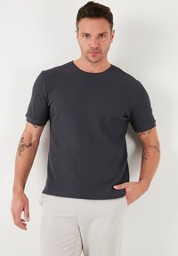 Tricou scurt cu mânecă, de culoare gri închis, cu un model texturat în relief și guler rotund, asociat cu pantaloni de culoare deschisă.