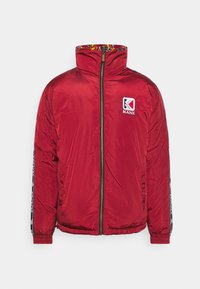 Červená prošívaná bunda s vysokým límcem, zapínáním na zip vpředu, elastickými manžetami a vzorovanou vnitřní podšívkou. Má logo na hrudi a kontrastní pruhy po stranách.