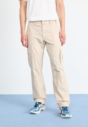 Beige cargo pants with multiple pockets, straight-leg design, and durable fabric. Worn with blue athletic shoes on a blue surface.