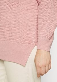 Różowy sweter dzianinowy z teksturowanym wzorem, ściągaczami przy mankietach i nieco dłuższym tylnym hemem. Noszony na jasnych spodniach.